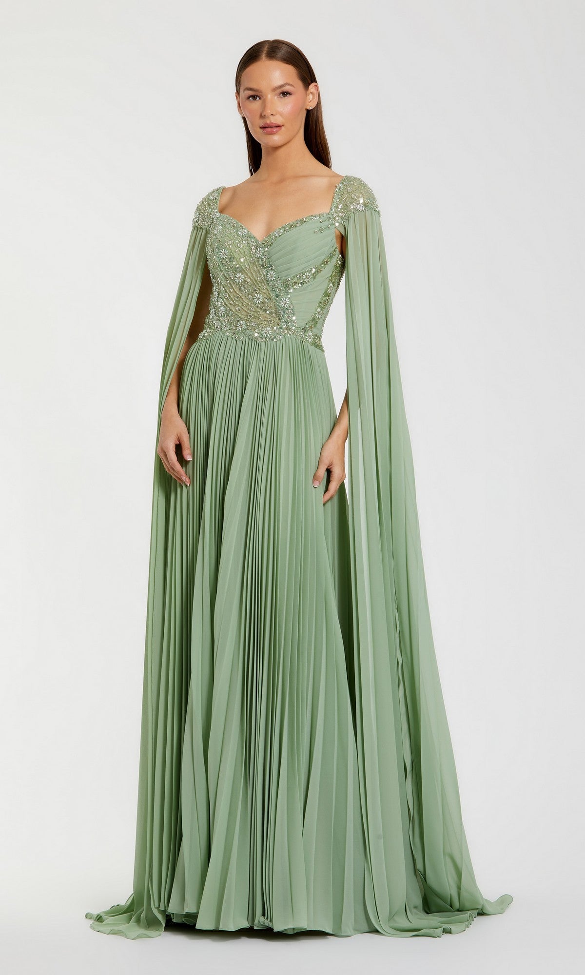 A woman poses in the Mac Duggal 37034 long formal dress in sage, featuring pleated chiffon, a floral-embellished bodice, and long draped cape sleeves against a plain background.